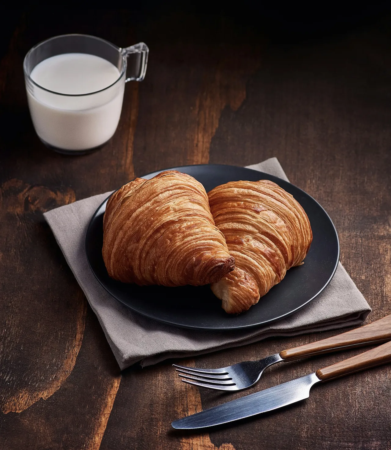 Dua croissant di piring gelap, segelas susu di sampingnya